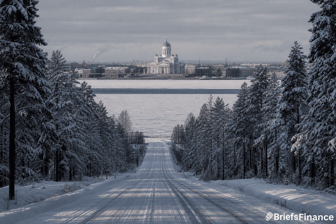 A snow-covered road lined with trees leads toward a frozen body of water and a city with a large white domed building in the background under a cloudy sky.