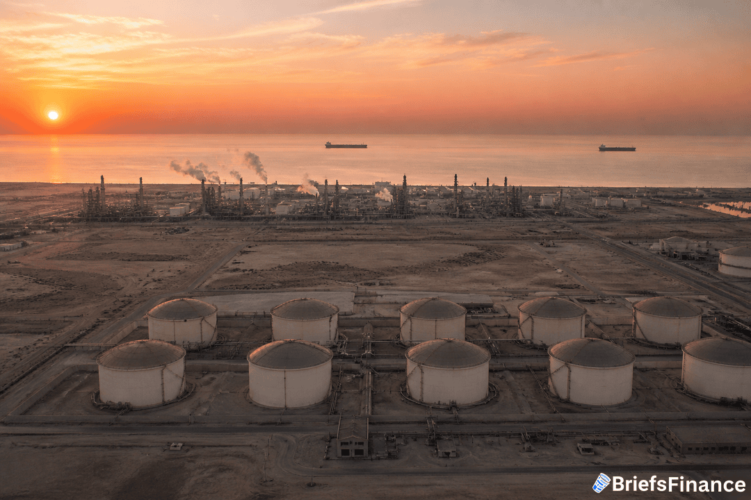 Aerial view of large oil storage tanks and refinery near the shore at sunset, with ships in the sea and smokestacks emitting vapor.