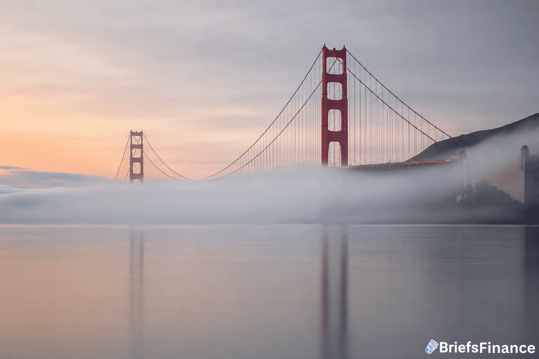 The Golden Gate Bridge is partially covered by fog during sunrise, with calm water in the foreground and hills in the background. The BriefsFinance logo is visible in the bottom right corner.