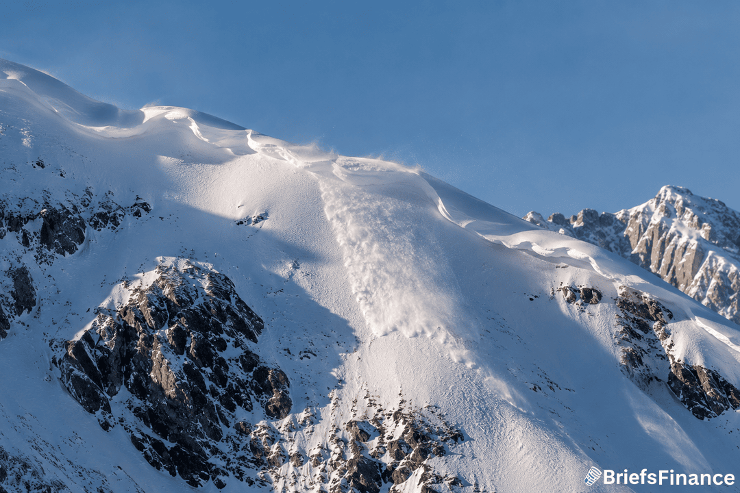 Snow pours down a rugged mountain slope during an avalanche, with wind blowing snow from the ridge under a clear blue sky. The BriefsFinance logo appears in the bottom right corner.