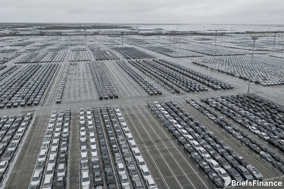 Aerial view of a vast parking lot filled with thousands of new cars arranged in neat rows under a cloudy sky, likely at a port or shipping facility.