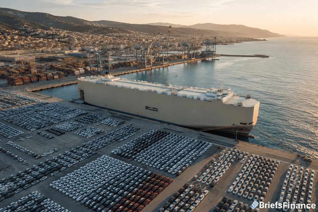 Aerial view of a large cargo ship docked at a port, with numerous rows of parked cars and shipping containers nearby. Cranes stand along the waterfront, and a coastal city with hills is visible in the background.