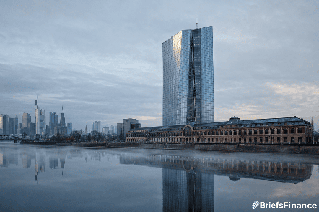 A modern glass skyscraper stands beside a historic building on a calm riverbank, reflecting in the water, with a city skyline and cloudy sky in the background. BriefsFinance logo appears in the bottom right corner.