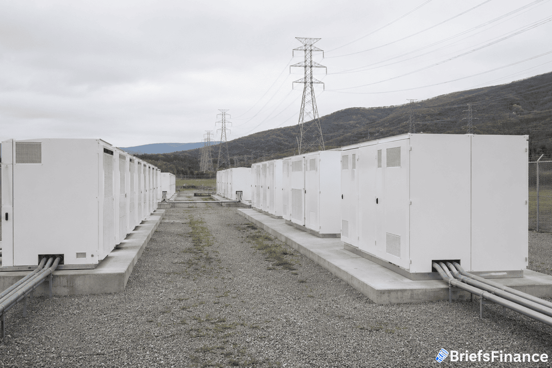Rows of large, white industrial battery storage units are installed outdoors on gravel, with power cables running into each unit. Power lines and hills are visible in the background under a cloudy sky.