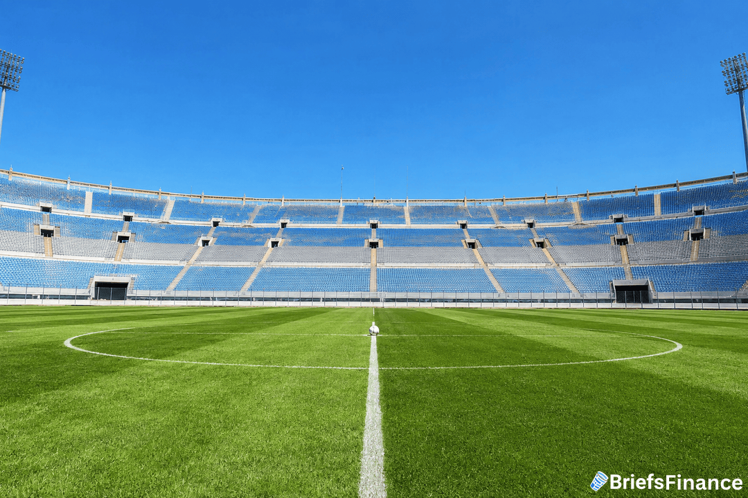 A large, empty soccer stadium with blue seats and bright floodlights under a clear blue sky. The view is from the center of the green grass field, with the BriefsFinance logo in the bottom right corner.